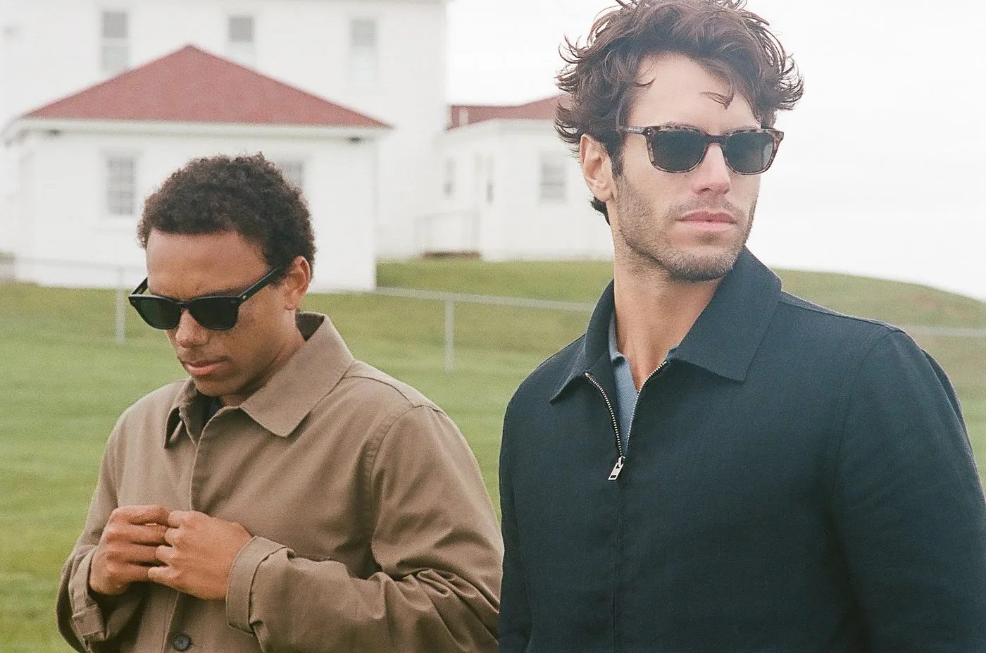 Two friends in sunglasses and jackets walking past a white shingled house on a coastal path